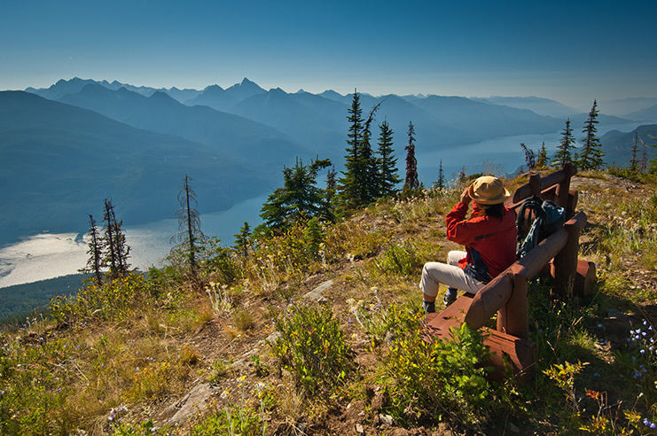 A person sitting on a hand-crafted wooden bench at the top of Mount Buchanan, looking down along Kootenay Lake