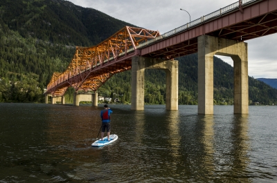 A man stand up paddle boarding under the Big Orange Bridge in Nelson, BC.