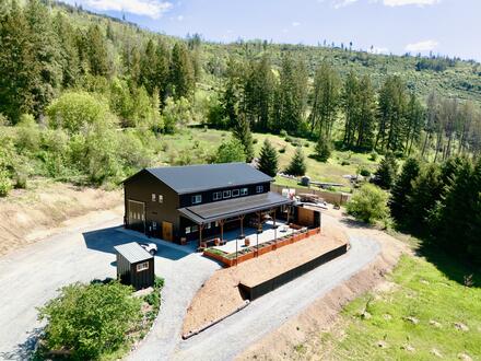 A large barn-style building with a black roof sits on cleared land surrounded by trees and hills, featuring a fenced patio and gravel driveway.