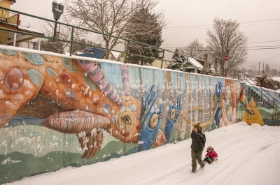 A mum pulling a child on a sled along the snow and next to a quirky mural in Nelson BC.