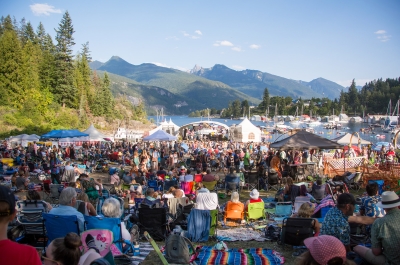 People sitting on the grass looking towards the floating stage on a sunny day at kaslo jazz festival