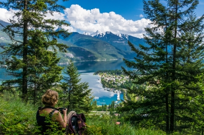 Woman with a camera sitting in the grass overlooking Kaslo, BC and Kootenay Lake
