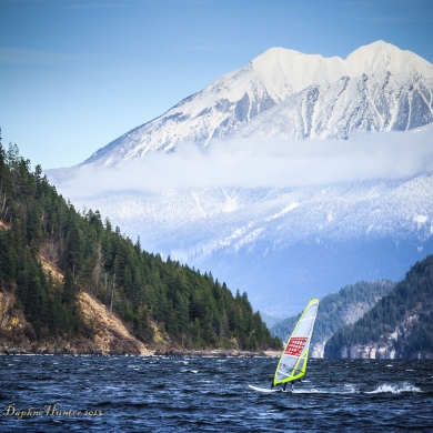 Windsurfer on Kootenay Lake, with treed mountainside and snow-covered mountain in the background