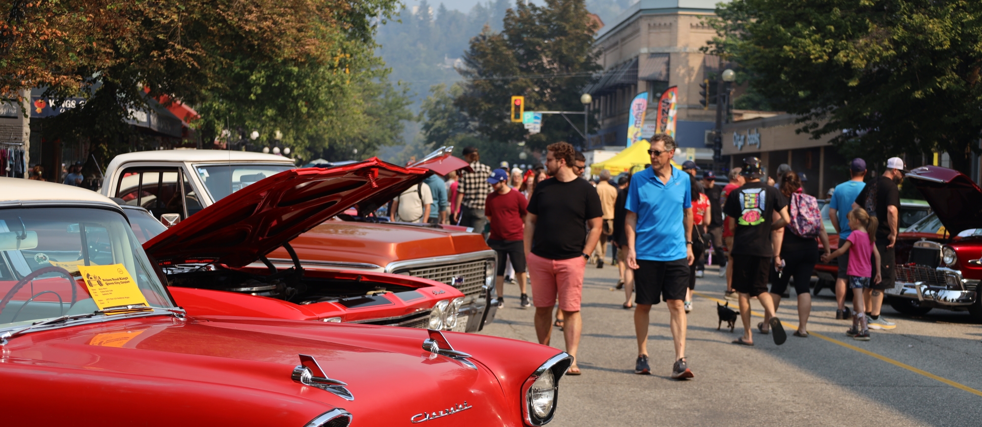 A red car in the foreground with people walking around enjoying the Queen City Car Show in Nelson. Photo by Janelle Lewchuk.