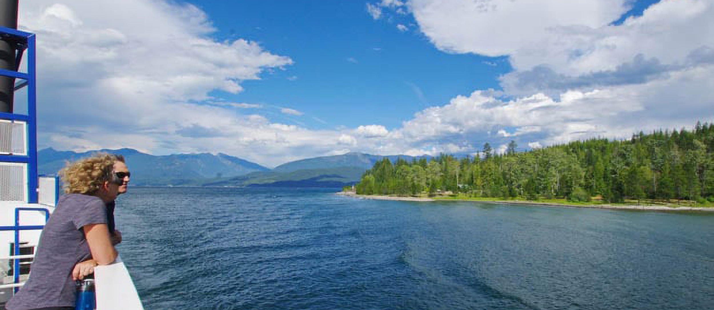 Two people on a ferry looking over the side at the lake on a sunny day, enjoying things to do near Nelson BC