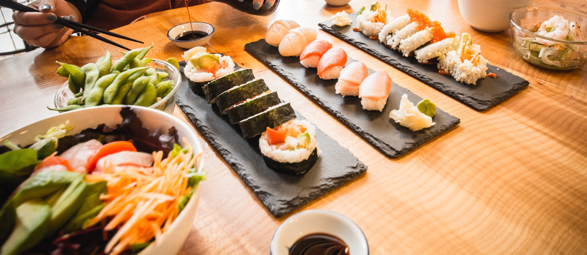 Hands pouirng soy sauce into a small dish with a table full of colourful sushi.