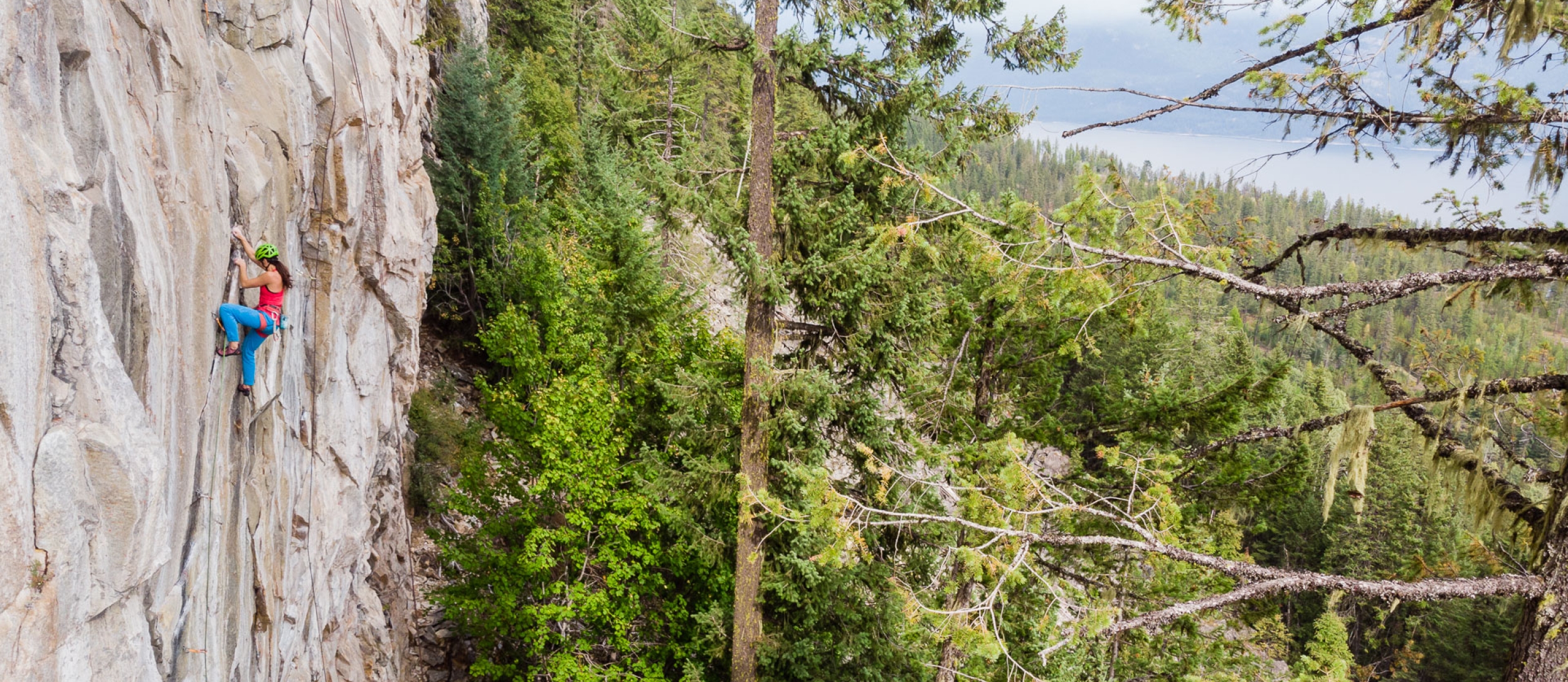 A rock climber on a steep wall with Kootenay Lake in the background.