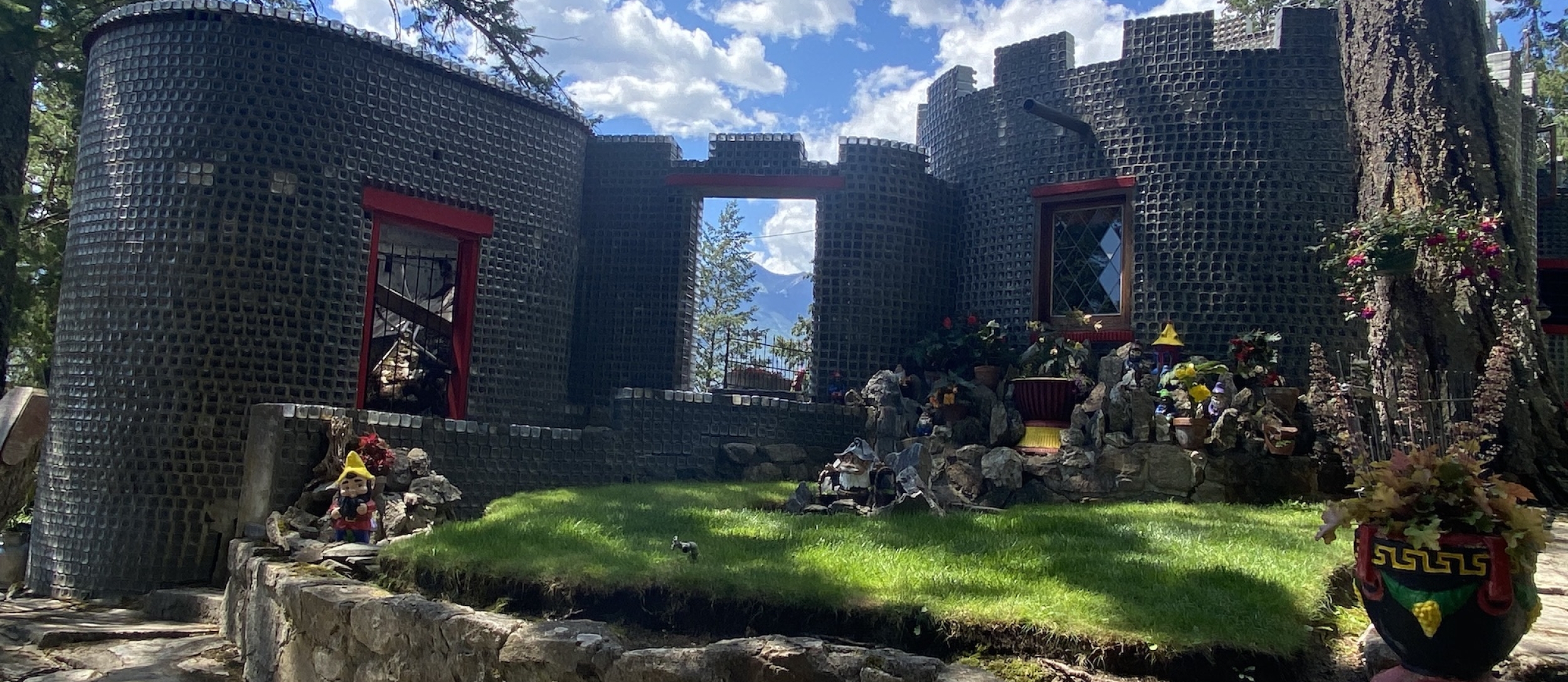 A castle-like house made out of glass bottles with green grass and lawn gnomes around it.