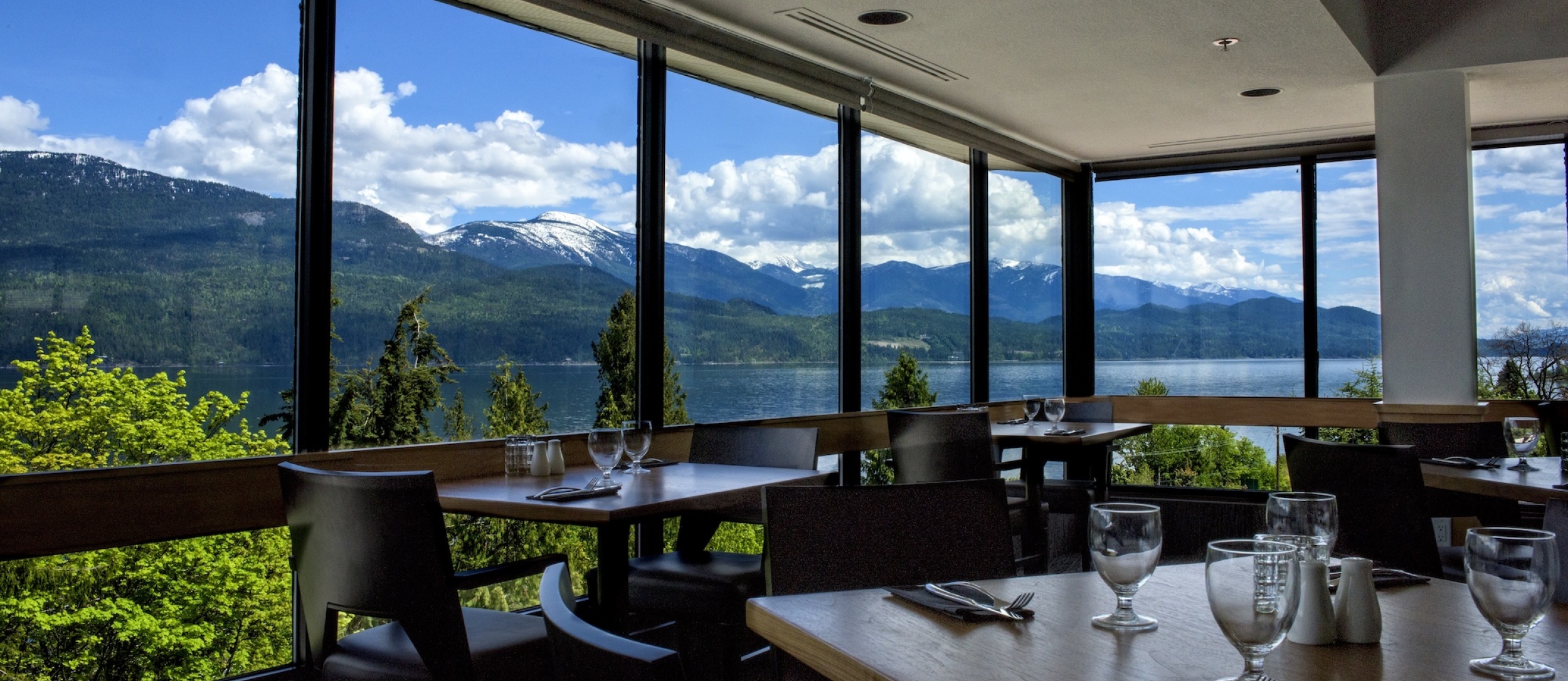 Fine dining tables which are set with wine glasses and cutlery. The sky and mountains can be seen through the glass windows