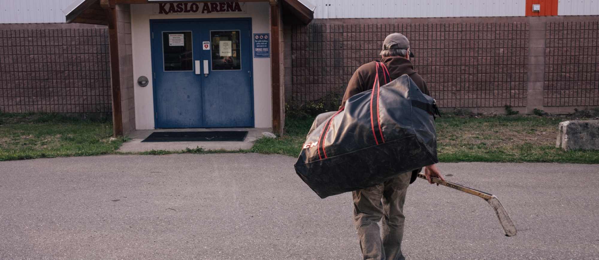 Man walking into Kaslo Arena holding a hockey stick and duffle bag