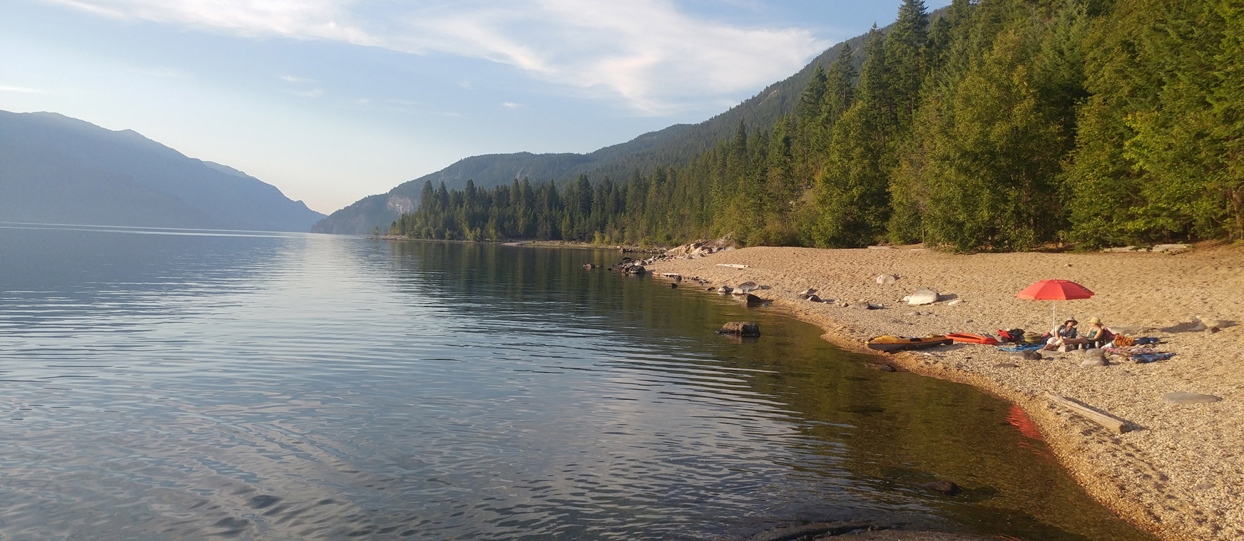 People sitting on a beach with a red sun umbrella at Barnard Beach near Riondel BC