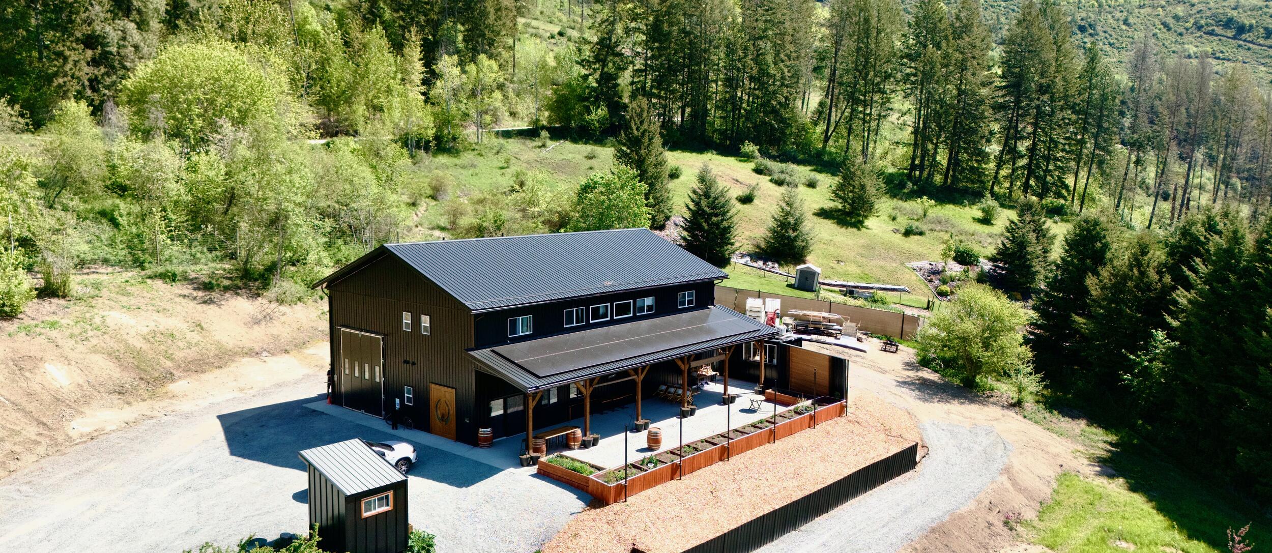 A large barn-style building with a black roof sits on cleared land surrounded by trees and hills, featuring a fenced patio and gravel driveway.