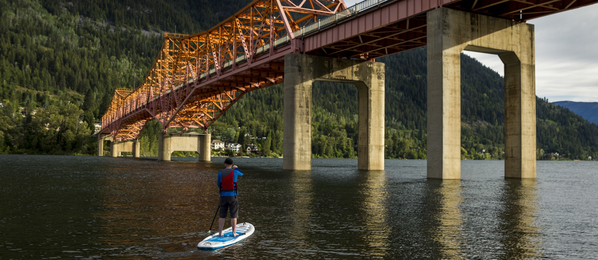 A man stand up paddle boarding under the Big Orange Bridge in Nelson, BC.