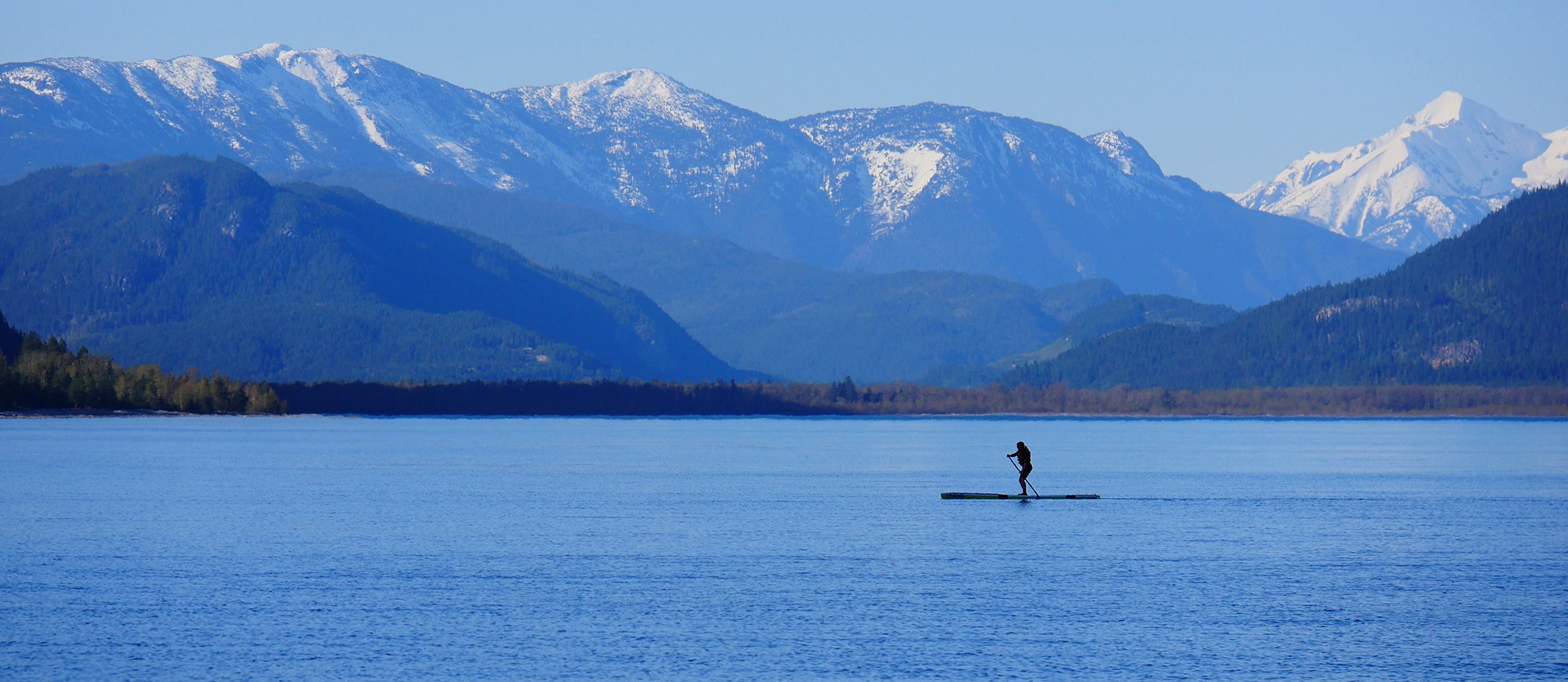 Silhouette of someone on a stand up paddle board in from of snow covered mountains.