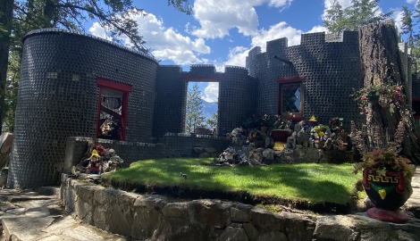 A castle-like house made out of glass bottles with green grass and lawn gnomes around it.