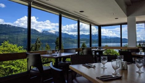 Fine dining tables which are set with wine glasses and cutlery. The sky and mountains can be seen through the glass windows