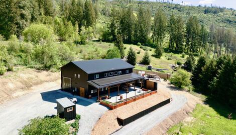 A large barn-style building with a black roof sits on cleared land surrounded by trees and hills, featuring a fenced patio and gravel driveway.