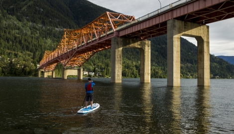 A man stand up paddle boarding under the Big Orange Bridge in Nelson, BC.